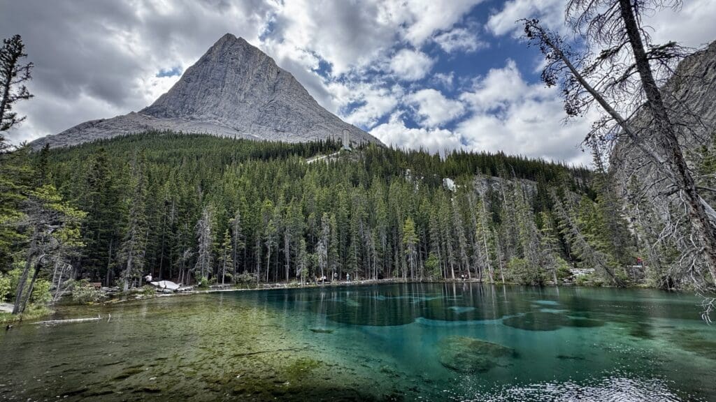 Majestic mountain under a cloudy sky, towering over a dense pine forest reflected in a tranquil turquoise Grassi Lakes in Kananaskis, Alberta