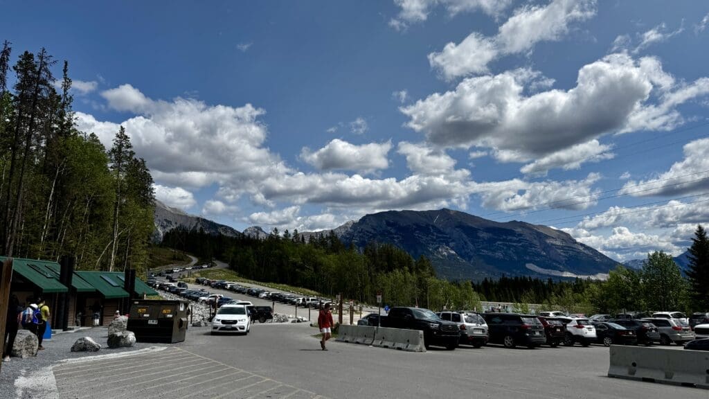 Mountain landscape with Grassi Lakes Trail parking lot in Kananaskis, Alberta surrounded by lush trees and a blue sky dotted with clouds.