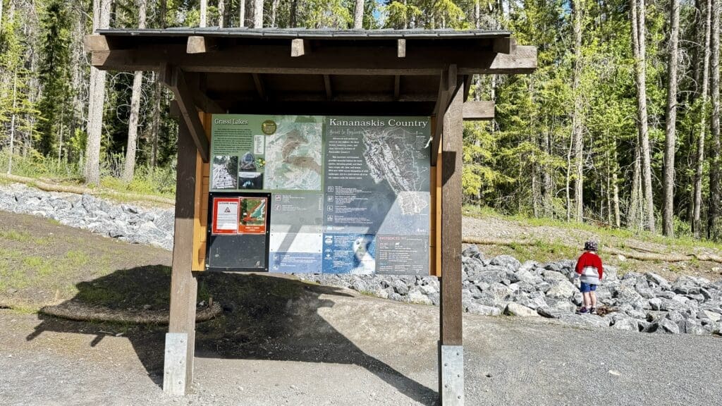 Wooden information board displaying maps and notices about Grassi Lakes and Kananaskis Country, surrounded by dense forest; a child in red stands nearby.