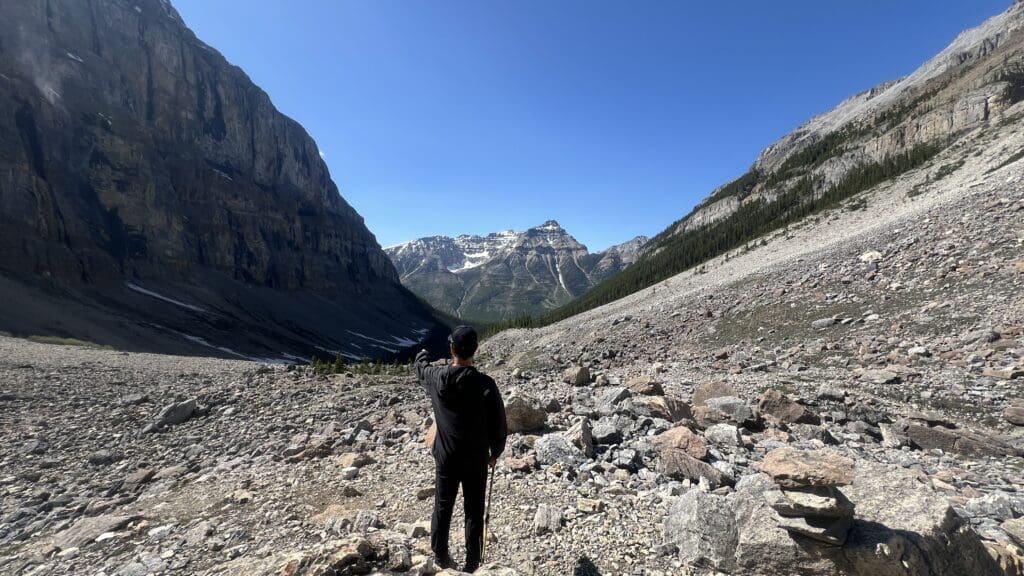 A person in black clothing stands on a rocky mountain path at Stanley Glacier Trail in Kootenay National Park, BC, pointing towards distant snow-capped peaks under a clear blue sky, conveying adventure.