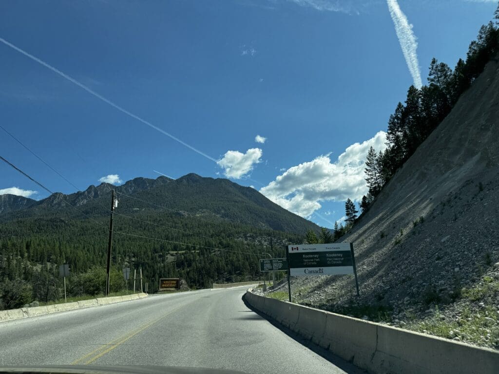 A winding road curves through a mountainous landscape under a clear blue sky with contrails. A sign marks the entrance to Kootenay National Park.