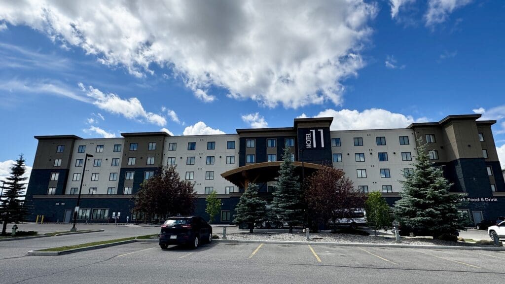 Four-story Hotel 11, MOD A Sonesta Collection in Calgary, Alberta under a partly cloudy sky. The facade is gray with a central wooden canopy. Trees and parked cars are in the foreground.