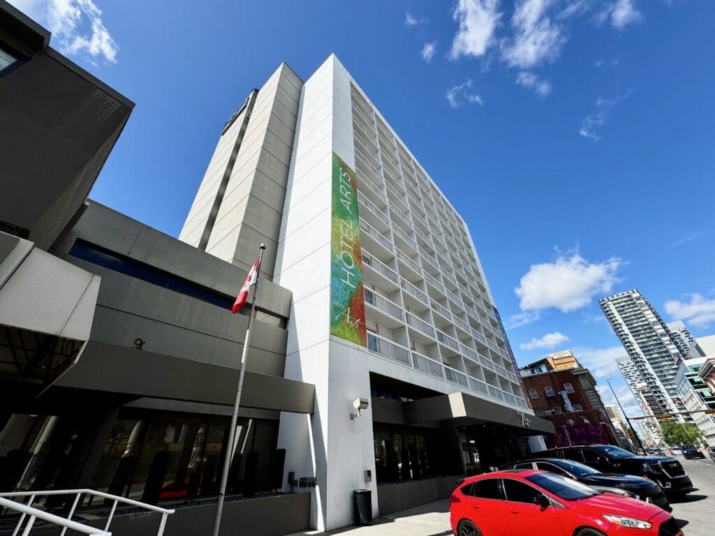 Tall white hotel in Calgary, Alberta with "Hotel Arts" banner, surrounded by a bright blue sky. A Canadian flag waves in front. Several parked cars, including a red one, on the street.