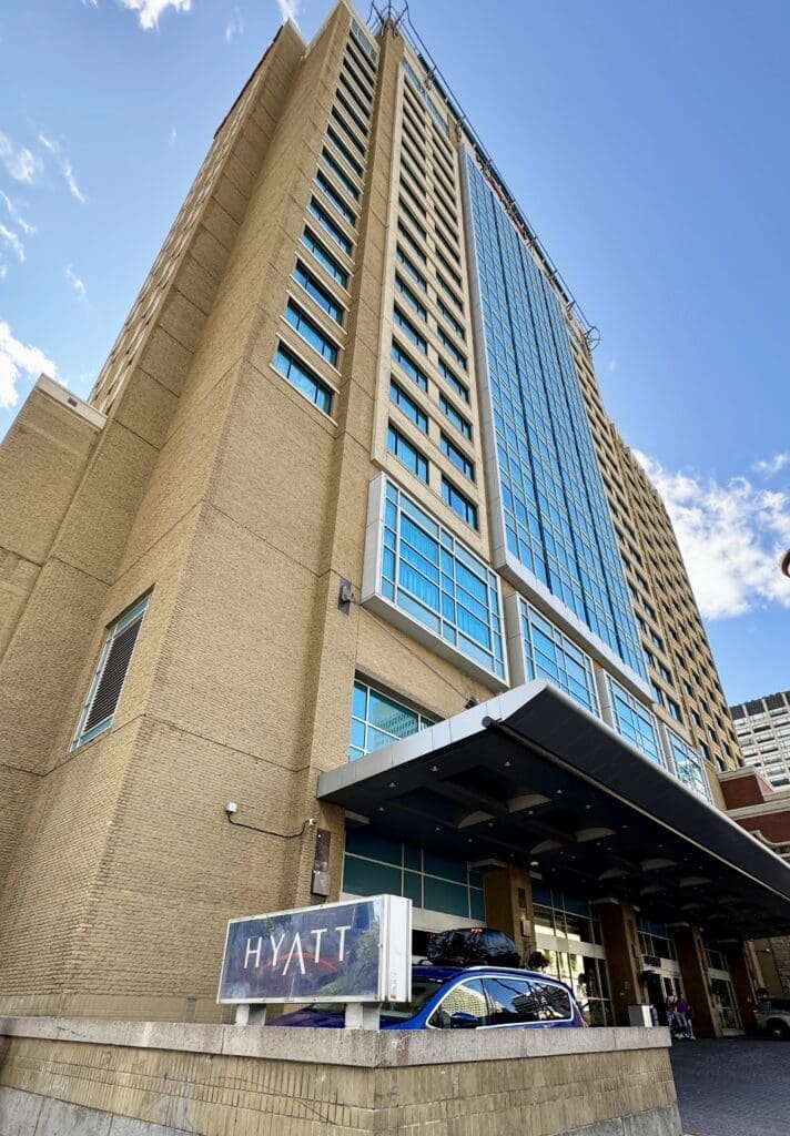 Tall beige Hyatt Regency in Calgary, Alberta with blue-tinted windows, viewed from below against a blue sky. A sign reads "Hyatt" near the entrance, conveying luxury.
