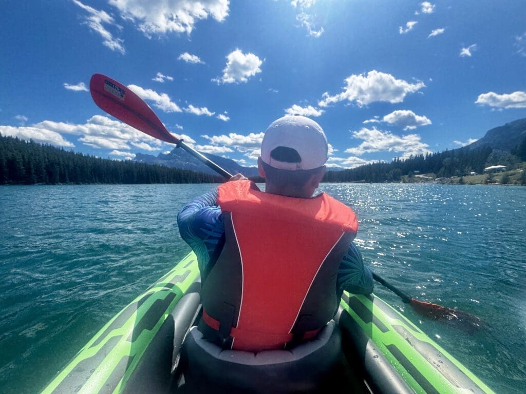 A person kayaking on a sparkling blue Two Jack Lake in Banff National Park, Alberta under a clear, sunny sky. They're wearing a red life jacket and a white cap, surrounded by forested mountains.