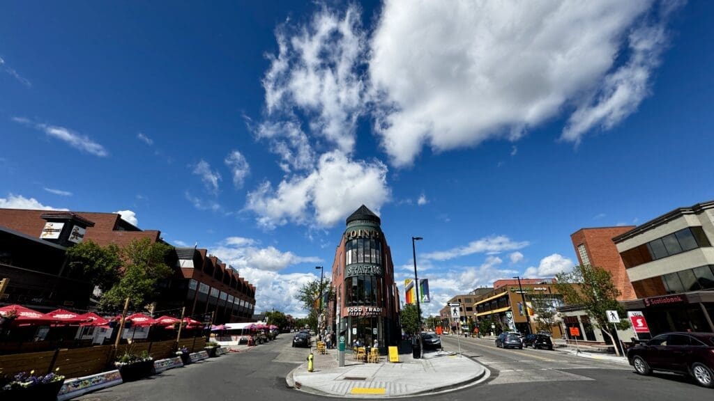 A bright, sunny street corner with a tall, rounded red-brick building labeled "Pointe" in the center located in Kensington in Calgary, Alberta. Red umbrellas line the left, and flags hang along the street. Fluffy clouds and a vivid blue sky enhance the lively urban scene.