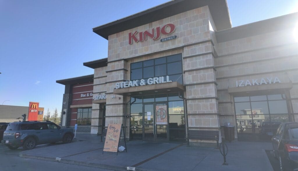 A restaurant named "Kinjo District" in Calgary, Alberta with a sign for "Steak & Grill" and "Izakaya" has a modern stone exterior. A car is parked nearby and there's a McDonald's in the distance. Sunlight creates a soft glare, evoking a bright, clear day.