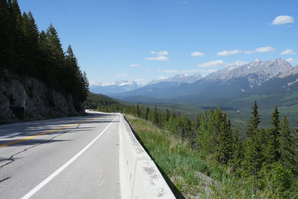 Mountain road with barrier on the left at Kootenay Valley Viewpoint in Kootenay National Park, BC, lined with dense trees. Snow-capped mountains stretch into the distance under a clear blue sky.