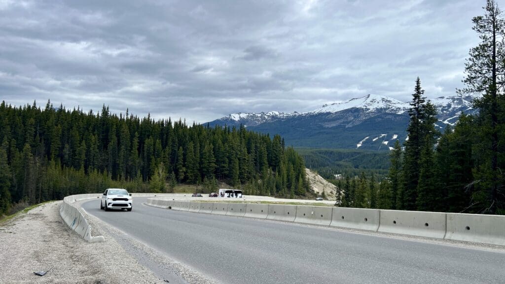 A white car drives on a curved mountain Lake Louise Drive in Banff National Park bordered by tall pine trees under a cloudy sky, with snow-capped mountains in the distance.