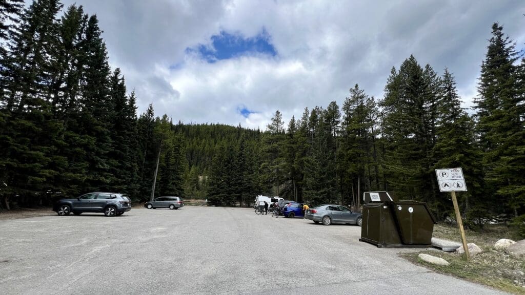 Parking lot surrounded by dense pine trees under cloudy skies with a patch of blue. Cars and a cyclist, sign, and bin in view; serene, natural setting.
