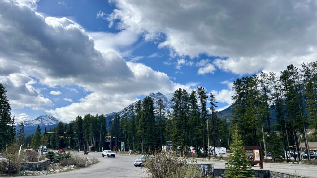 Mountain landscape in Lake Louise Village in Banff National Park with snow-capped peaks under a cloudy sky. Tall pine trees line a road with cars, capturing a serene and grand atmosphere.