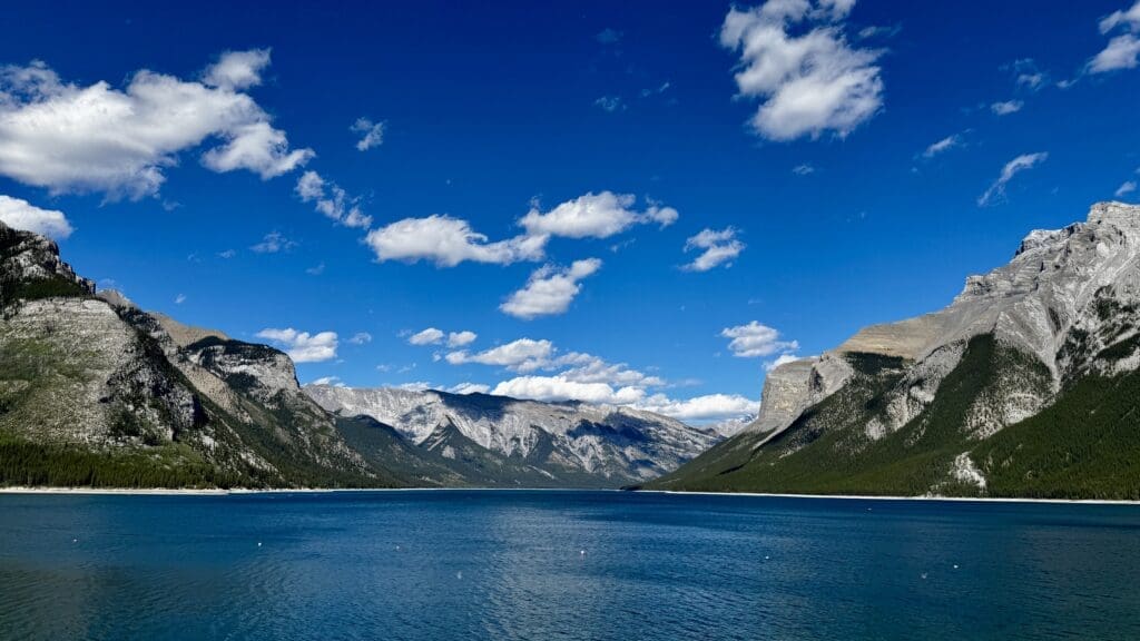 Lake Minnewanka in Banff National Park, Alberta under a vibrant blue sky, surrounded by towering mountains. The scene conveys a serene and majestic natural beauty.