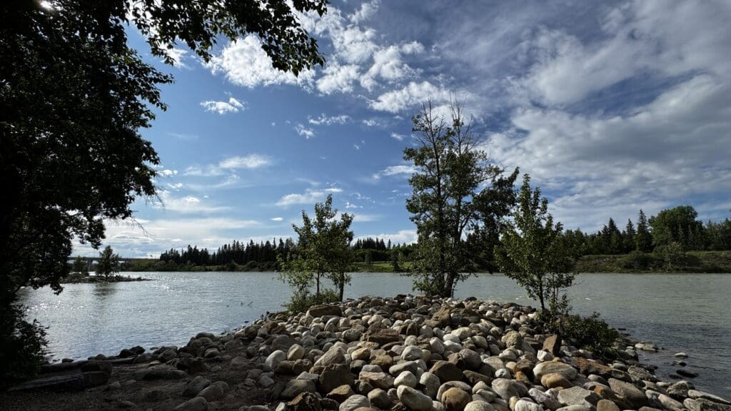 Rocky shoreline in Bowness Park in Calgary, Alberta with scattered trees by Bow River on a clear day. The sky is bright blue with fluffy clouds, evoking a calm and serene atmosphere.
