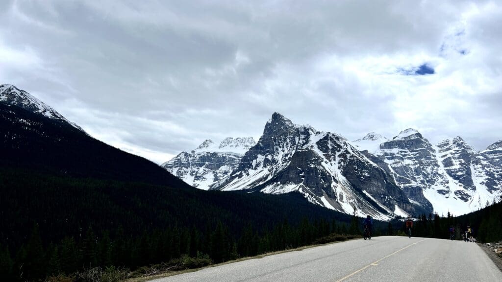 A snowy mountain range under a cloudy sky, with cyclists riding along Moraine Lake Road in the foreground.