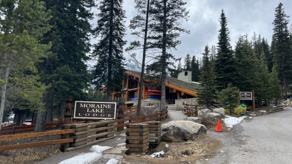 Mountain lodge surrounded by tall pine trees with a sign reading “Moraine Lake Lodge.” Canoes are stacked outside the rustic wooden building, set against a cloudy sky.