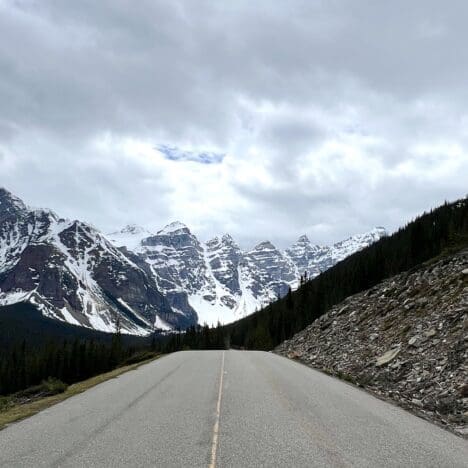 The deserted Moraine Lake Road stretches toward snow-capped mountains under a cloudy sky. Pine trees line the road, creating a serene and majestic landscape.