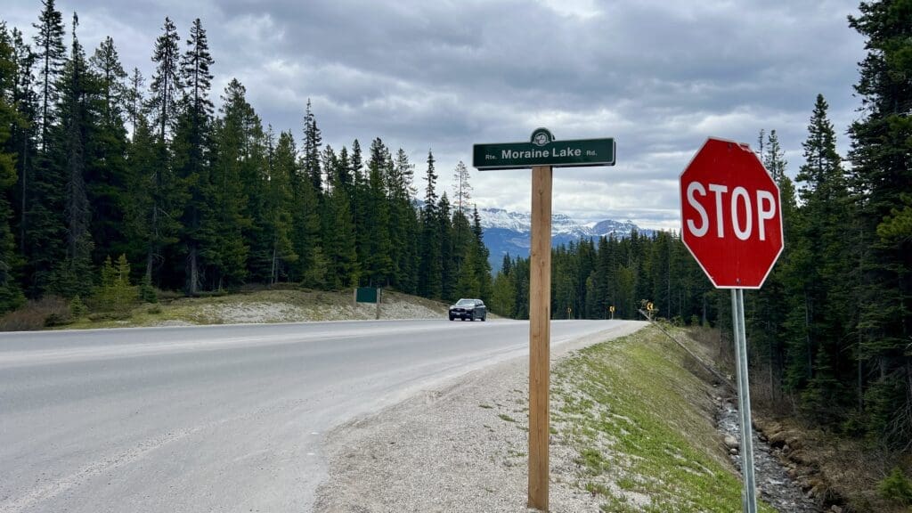 Road scene in Banff National Park with a "Moraine Lake Rd" sign and stop sign, surrounded by tall evergreen trees. A car approaches on a cloudy day, mountains in the background.