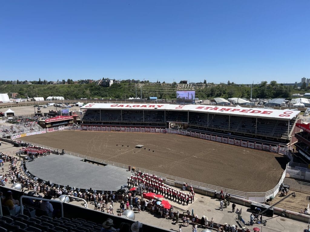 Arial view of a large outdoor arena at the Calgary Stampede, with grandstands and dirt showground. Crowd gathers near the entrance under a clear blue sky.