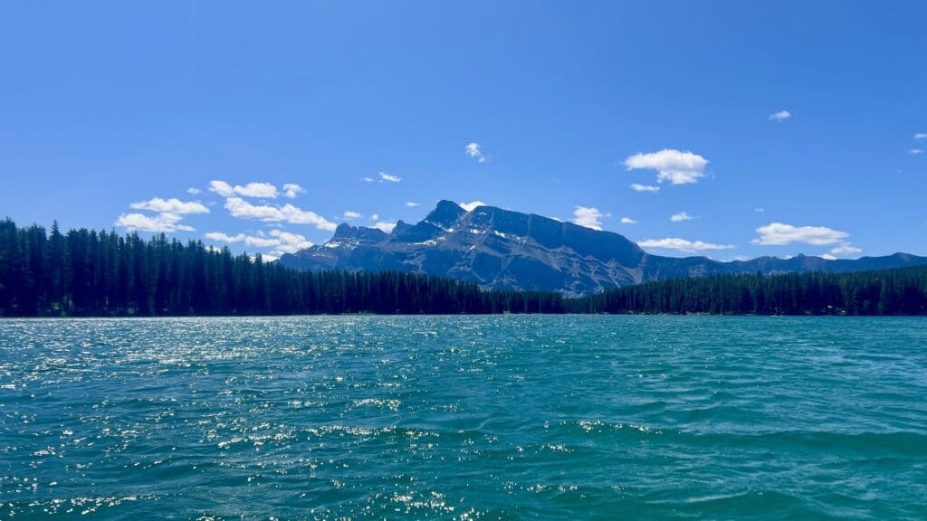 Two Jack Lake in Banff National Park, Alberta with sparkling blue water is surrounded by dense pine forests. Majestic Mount Rundle tower in the background under a clear, sunny sky.