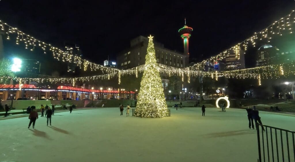 A festive ice rink in Olympic Plaza in Calgary, Alberta with a large, illuminated Christmas tree in the center. Skaters glide around, surrounded by strings of glowing lights at night.