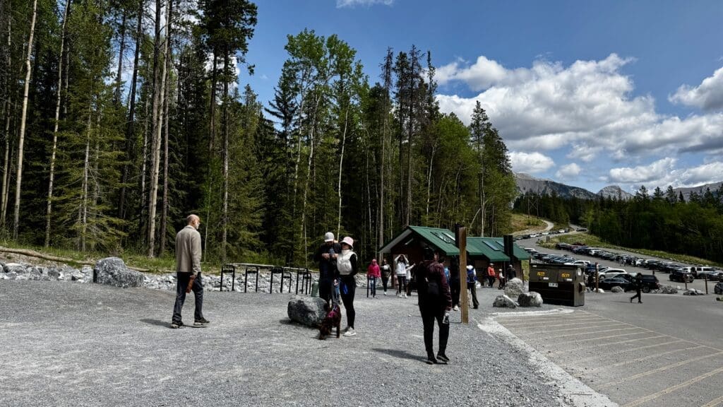 A wooded area with tall pine trees surrounds Grassi Lakes parking lot in Kananaskis, Alberta and building with a green roof. People in jackets walk on a gravel path under a partly cloudy sky.