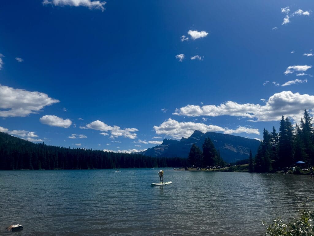 Two Jack Lake in Banff National Park, Alberta with a person paddleboarding under a clear blue sky. Evergreen trees surround the water, and distant mountains enhance the tranquil scene.