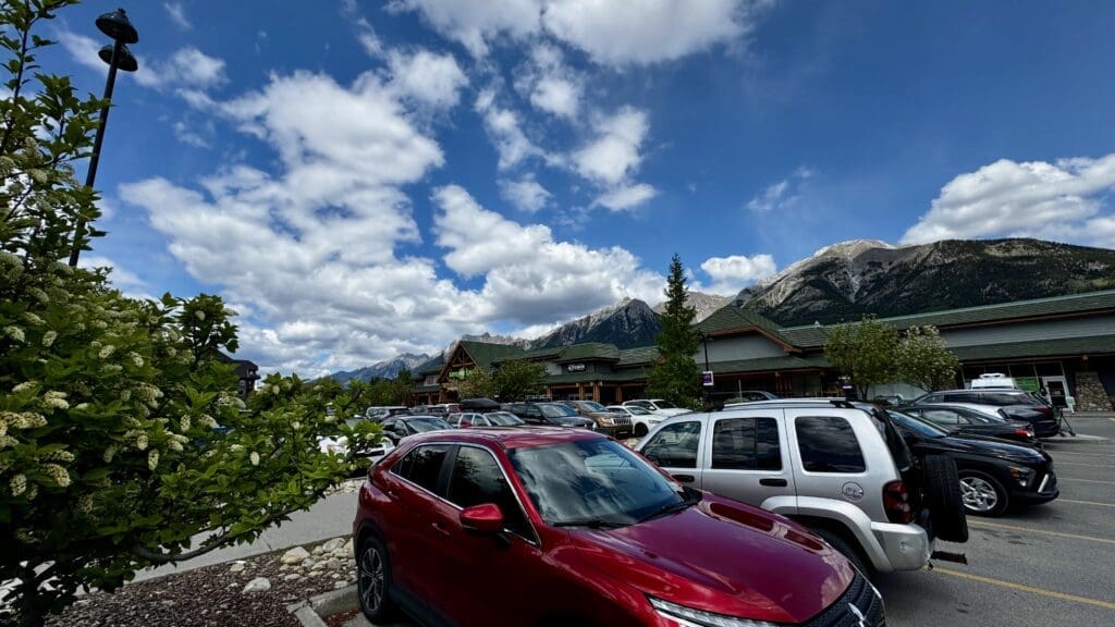 Clear blue sky with fluffy clouds over a parking lot in Canmore Town, Alberta filled with cars, a red SUV prominent. Mountains and shops in the background, greenery to the left.