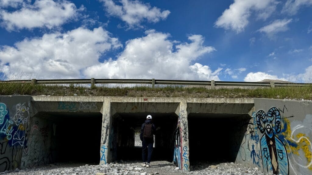 A person in a white cap walks into a graffiti-decorated underpass beneath a road, towards Three Sisters Viewpopint in Canmore, Alberta, against a bright blue sky with scattered clouds. Rugged and artistic.