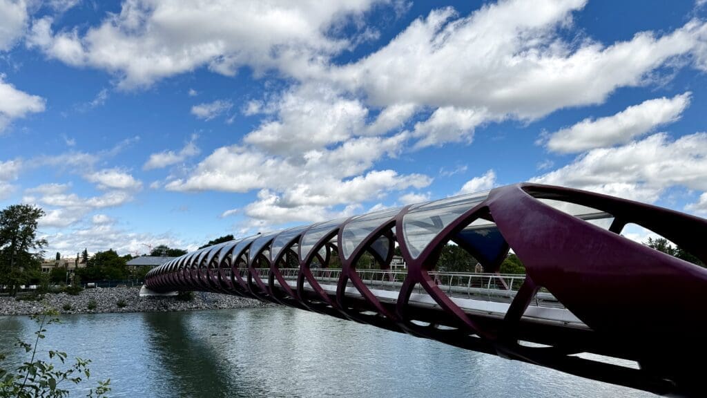 Red tubular Peace Bridge in Calgary, Alberta with diamond-patterned supports spans a calm Bow River under a vibrant blue sky with scattered clouds, conveying a modern feel.