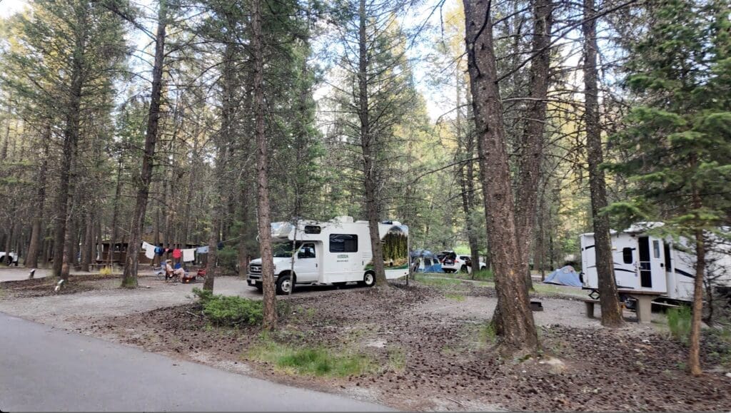 RV and tents in Redstreak Campground in Radium, BC. A family sits by a fire with laundry hanging between trees. Peaceful, natural setting with tall pine trees.
