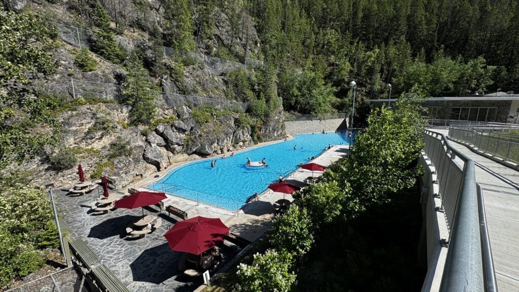 Radium Hot Springs in Kootenay National Park, BC is a scenic outdoor pool is nestled against a rocky hillside surrounded by lush trees. Several people swim and relax under red umbrellas on a sunny day.