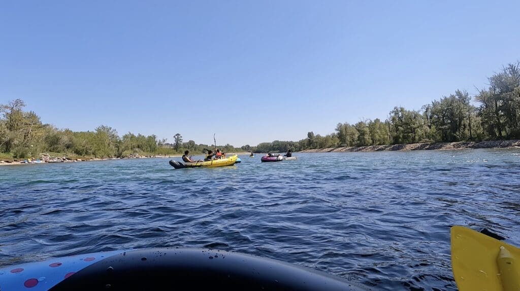 People in colorful inflatable rafts paddle on Bow River in Calgary, Alberta under a clear blue sky, surrounded by lush green trees. The scene is lively and adventurous.