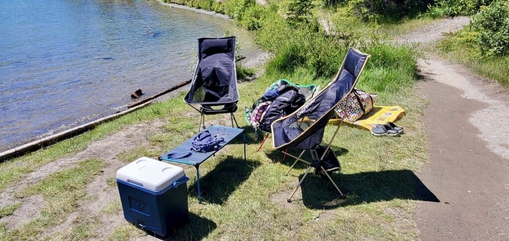 Camping setup by Two Jack Lake in Banff National Park, Alberta includes two chairs, a table with a hat, cooler, colorful bags, and a mat on grass. Sunny, serene outdoor scene.