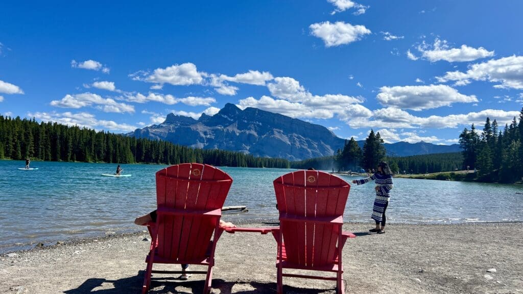Two red Adirondack chairs at Two Jack Lake in Banff National Park, Alberta face a tranquil lake with paddleboarders. Mountains and pine trees enhance the serene, sunny scene.