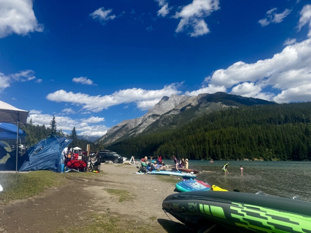 A scenic lakeside view at Two Jack Lake in Banff National Park, Alberta on a sunny day, with people enjoying the water and beach. Mountains and pine forests are visible under a vibrant blue sky.