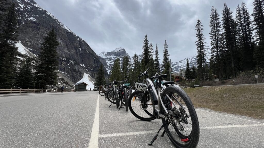 Row of parked bicycles on Moraine Lake Parking Lot in Banff National Park, with snow-capped peaks and tall pine trees in the background under a cloudy sky, conveying a serene outdoor adventure mood.