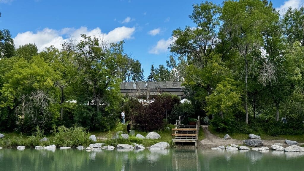 Calm riverbank scene with lush green trees and a sign for River Café in Calgary, Alberta. A wooden path leads down to the water, conveying a peaceful, inviting atmosphere.