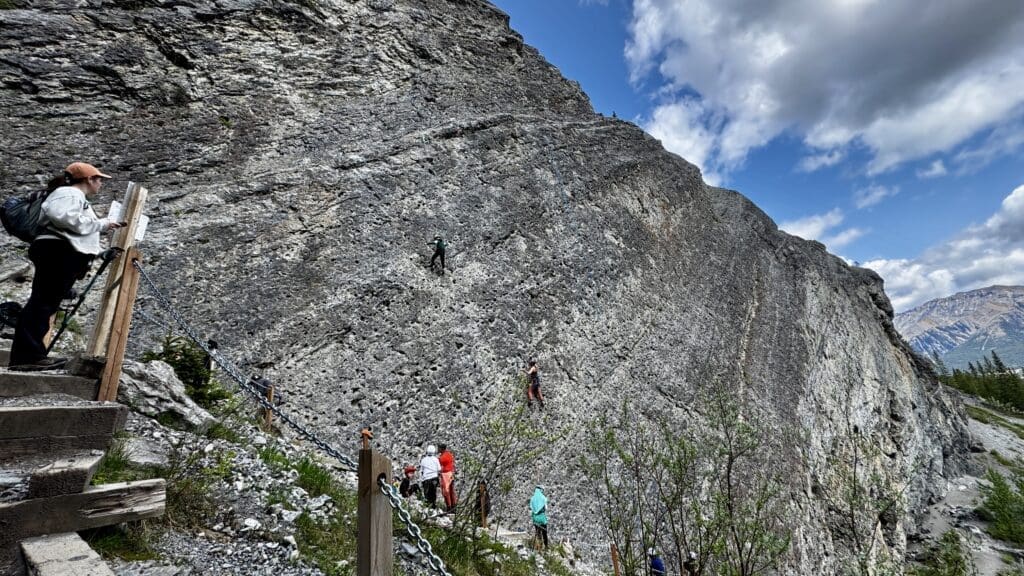 A group of climbers scale a steep rocky cliff under a partly cloudy blue sky. A person observes from wooden steps, surrounded by lush greenery. Energetic and adventurous.
