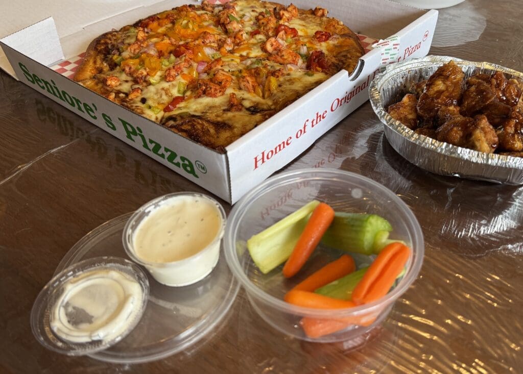 A pizza box of Seniore's Pizza in Calgary, Alberta sits open, revealing a colorful pizza with peppers and chicken. Nearby are saucy chicken wings, ranch dip, and a cup of carrot and celery sticks.