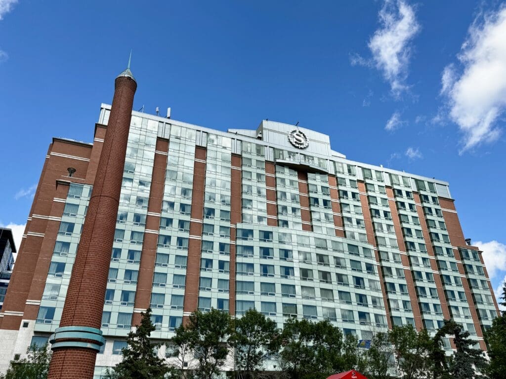 Tall brick and glass Sheraton Suites Calgary Eau Claire in Calgary, Alberta under a bright blue sky, with a prominent chimney-like structure in the foreground and green trees at the base.