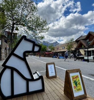 Outdoor street scene in Canmore Town, Alberta with a geometric polar bear sculpture on a wooden platform. People stroll past rustic buildings under a cloudy sky, with mountain peaks in the background.