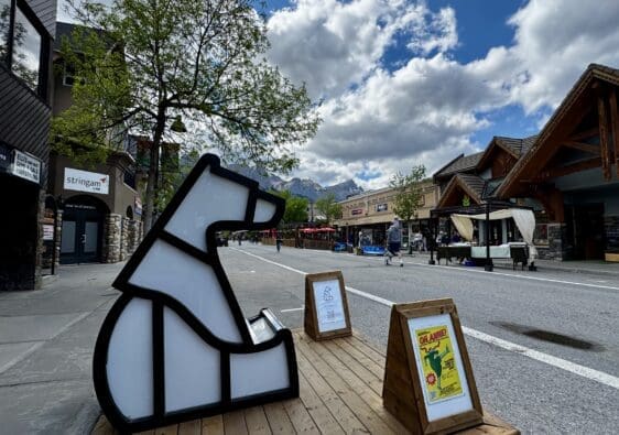 Outdoor street scene in Canmore Town, Alberta with a geometric polar bear sculpture on a wooden platform. People stroll past rustic buildings under a cloudy sky, with mountain peaks in the background.