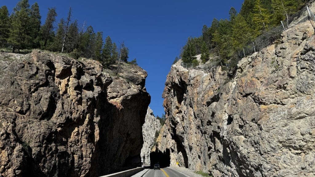 A car drives through the narrow Sinclair Canyon in Kootenay National Park, BC with towering, rugged rock walls under a clear blue sky. Tall trees line the cliff edges, adding natural beauty.