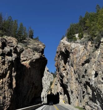 A car drives through the narrow Sinclair Canyon in Kootenay National Park, BC with towering, rugged rock walls under a clear blue sky. Tall trees line the cliff edges, adding natural beauty.