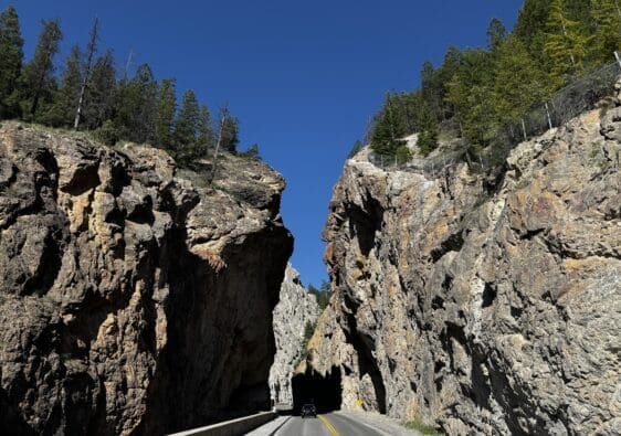 A car drives through the narrow Sinclair Canyon in Kootenay National Park, BC with towering, rugged rock walls under a clear blue sky. Tall trees line the cliff edges, adding natural beauty.