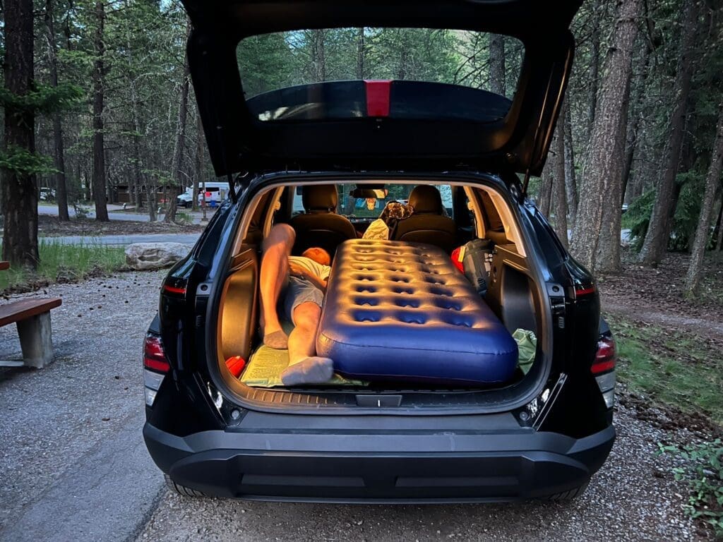 Open trunk of a black SUV with a blue air mattress inside. A person is lying on the mattress. Surrounding area is the wooded Redstreak Campground in Radium, BC with a rustic, peaceful vibe. camping essentials checklist