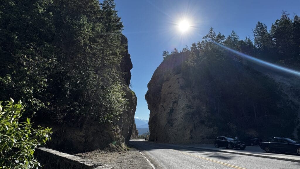 Sunlit mountain pass with tall, rocky cliffs of Sinclair Canyon in Kootenay National Park, BC on either side. A paved road curves through, bordered by dense, green forest. Cars park under a bright, clear sky.