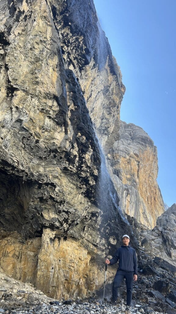 Man in outdoor gear stands by a large rocky mountain at Stanley Glacier Trail in Kootenay National Park, BC with a trickling waterfall, under a clear blue sky. The scene conveys adventure and tranquility.