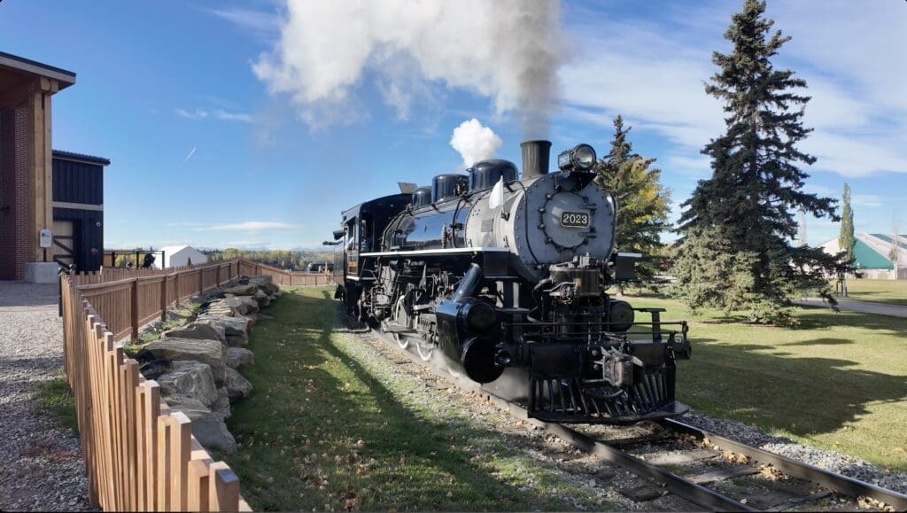 A black steam locomotive numbered 2023 at Heritage in Calgary, Alberta emits white smoke under a vibrant blue sky. Surrounded by trees, it runs past a wooden fence and a brick building, conveying nostalgia.