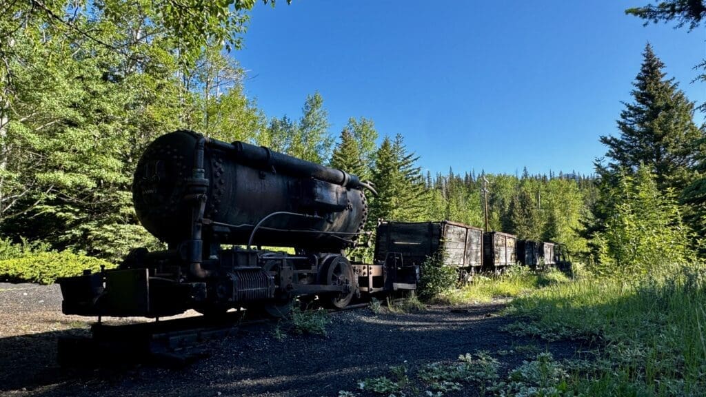 Old, rusted train cars at Bankhead Loop in Banff National Park, Alberta sit on tracks surrounded by lush green forest under a bright blue sky. The scene feels serene and abandoned.